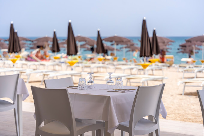Tavolo apparecchiato, tovaglia e sedie bianche davanti alla spiaggia con lettini, ombrelloni chiusi e mare azzurro