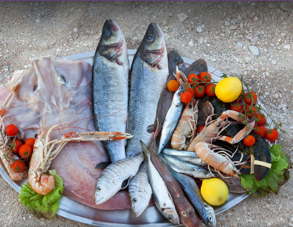 Vassoio d'acciaio con branzini, razza, scampi, sgombri, aragosta, pomodorini, limoni e insalata appoggiato su sabbia e ciottoli